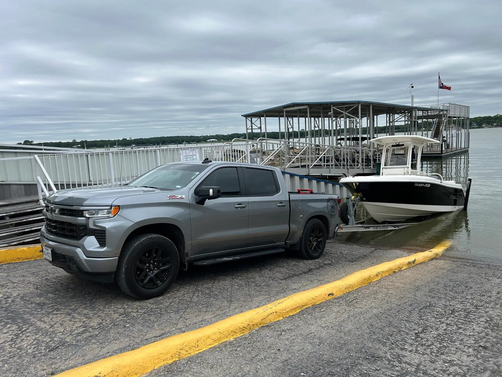 A silver pickup truck is parked at a boat ramp, towing a white and black boat partially in the water. The sky is overcast, and a small dock with a covered structure is visible in the background.